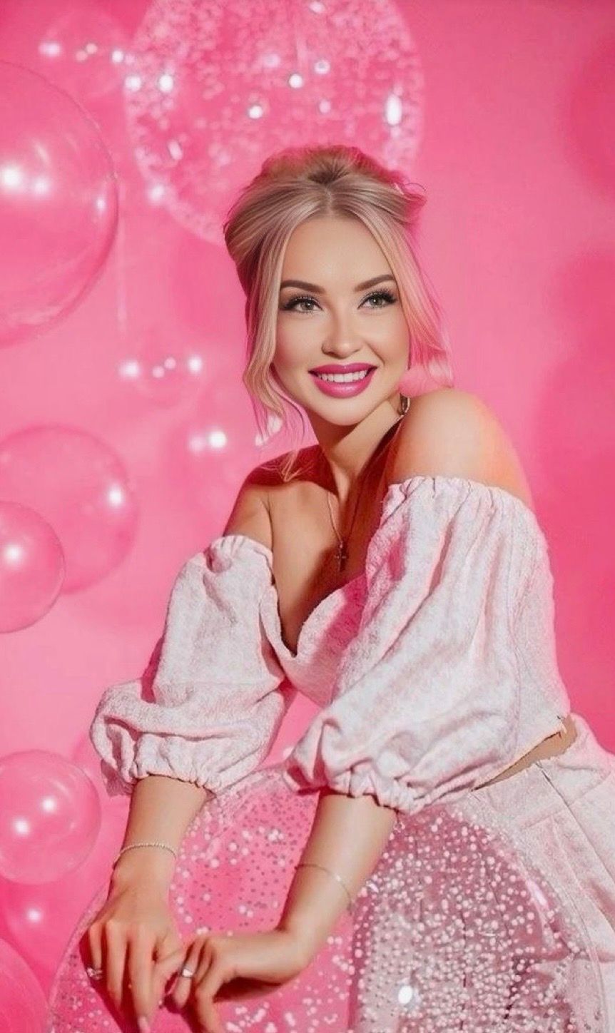A smiling blonde woman in an off-shoulder pink outfit poses against a vibrant pink background with shimmering balloons.