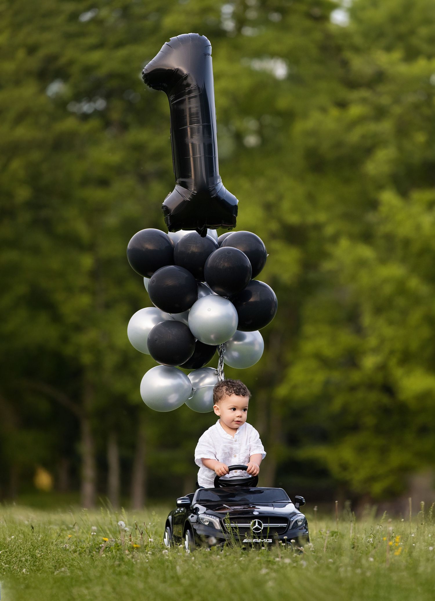 Baby boy sits on toy Mercedes car with black and silver number 1 balloons in park for first birthday.