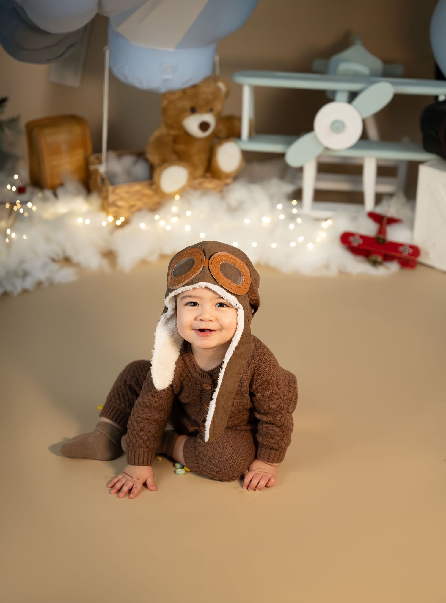 Smiling baby in brown knit aviator outfit and pilot hat sits on beige floor with teddy bear and toy airplane backdrop.