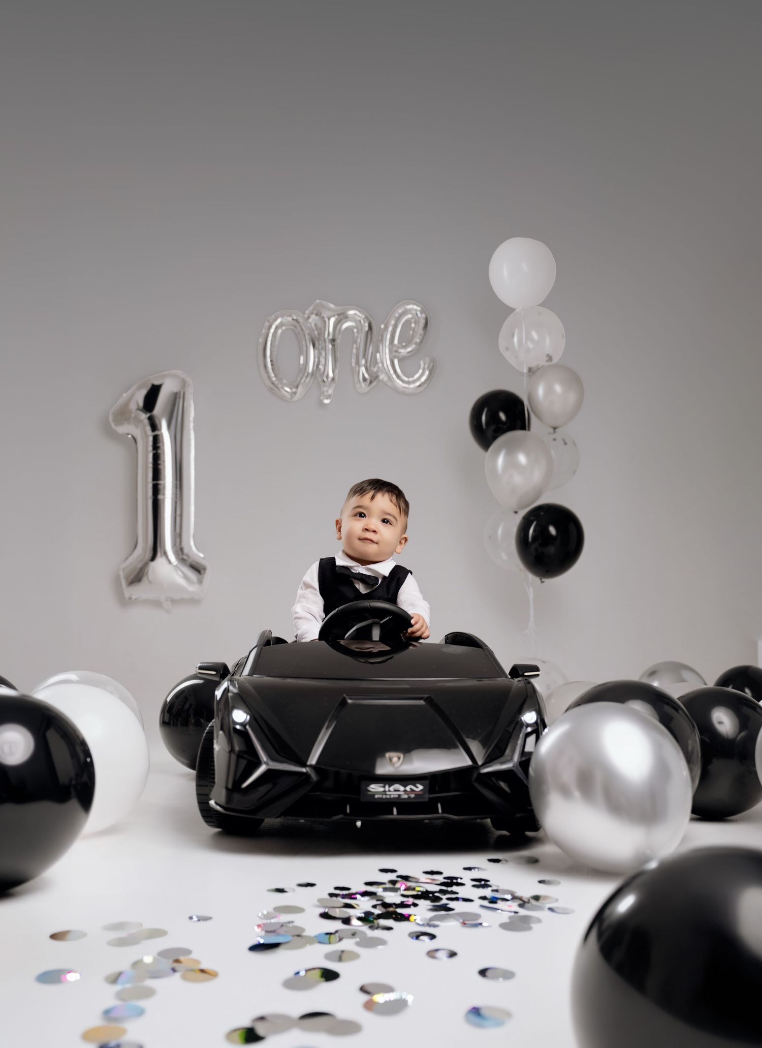 Baby boy in suit sits in black toy Lamborghini surrounded by black and silver balloons celebrating his first birthday.