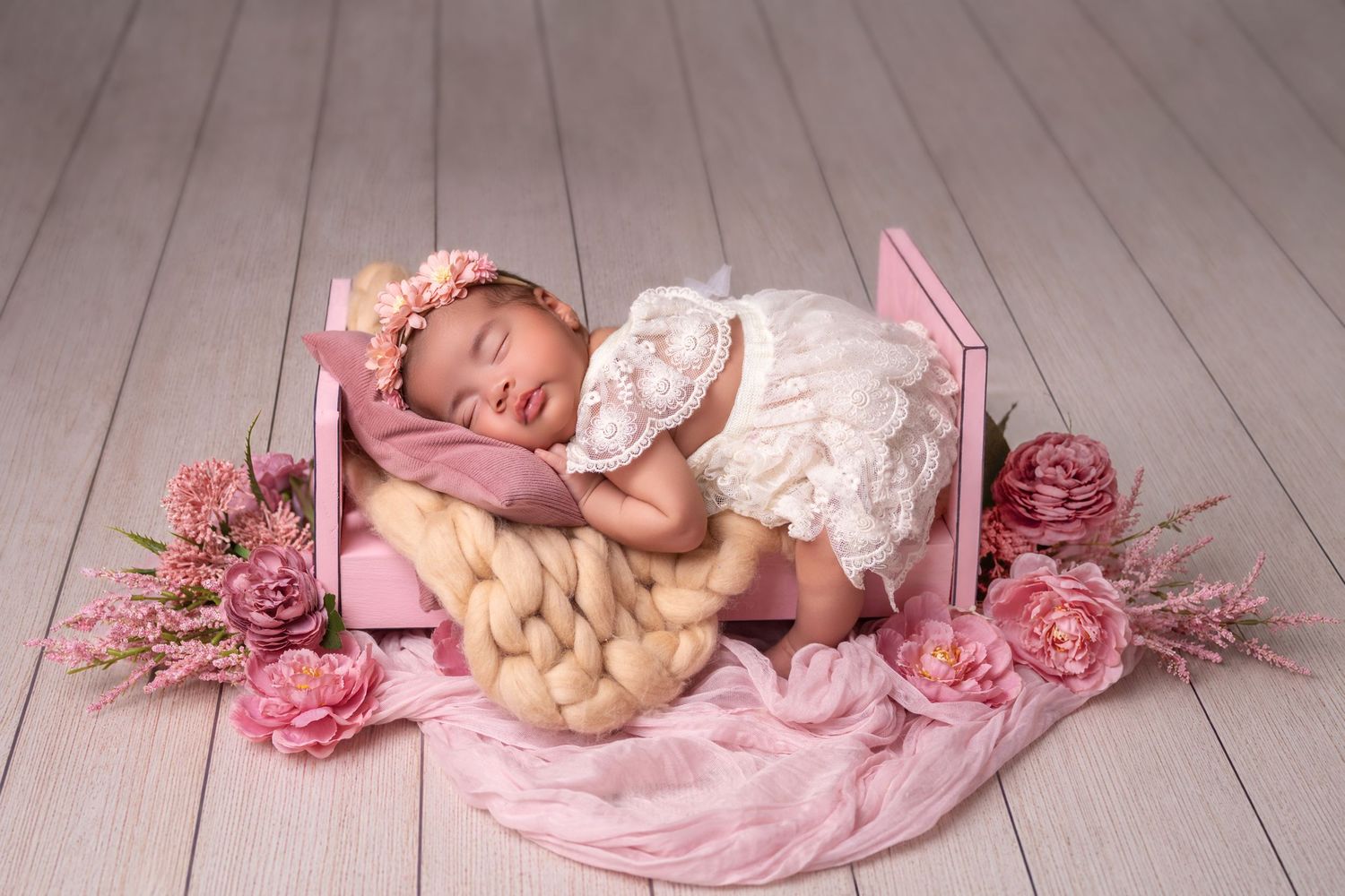 Newborn baby sleeping in a tiny pink wooden bed surrounded by pink roses and a chunky knit blanket.