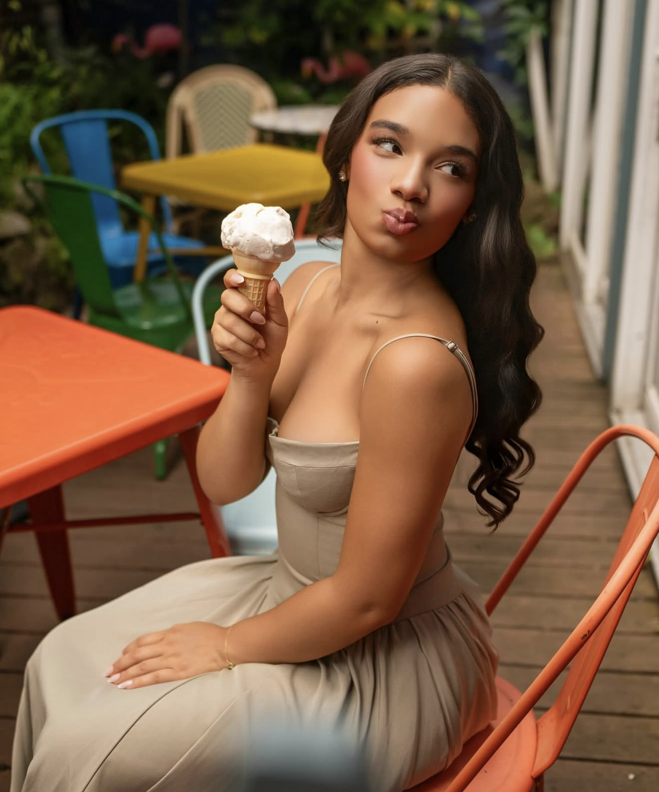 Young woman with long wavy hair sitting outdoors holding an ice cream cone, wearing a beige dress at a colorful cafe.