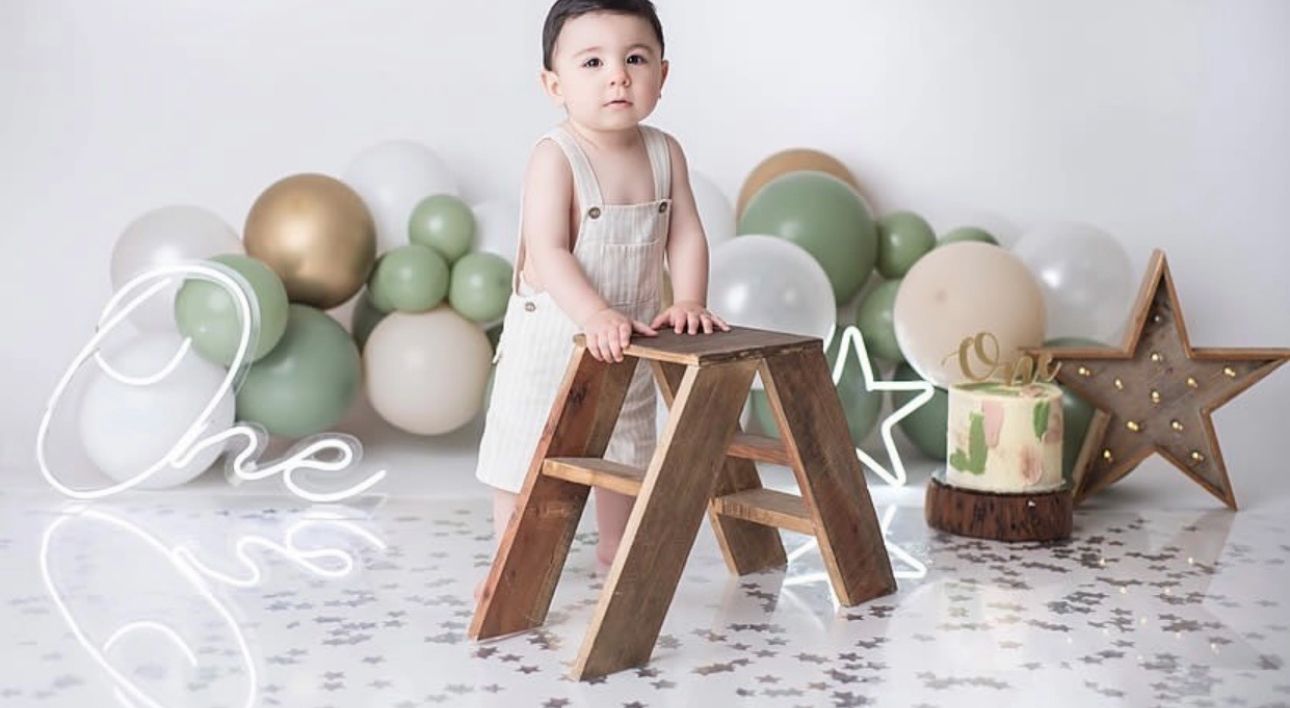 A small child sits atop a wooden ladder surrounded by sage green and gold balloons with a decorative star in the background.