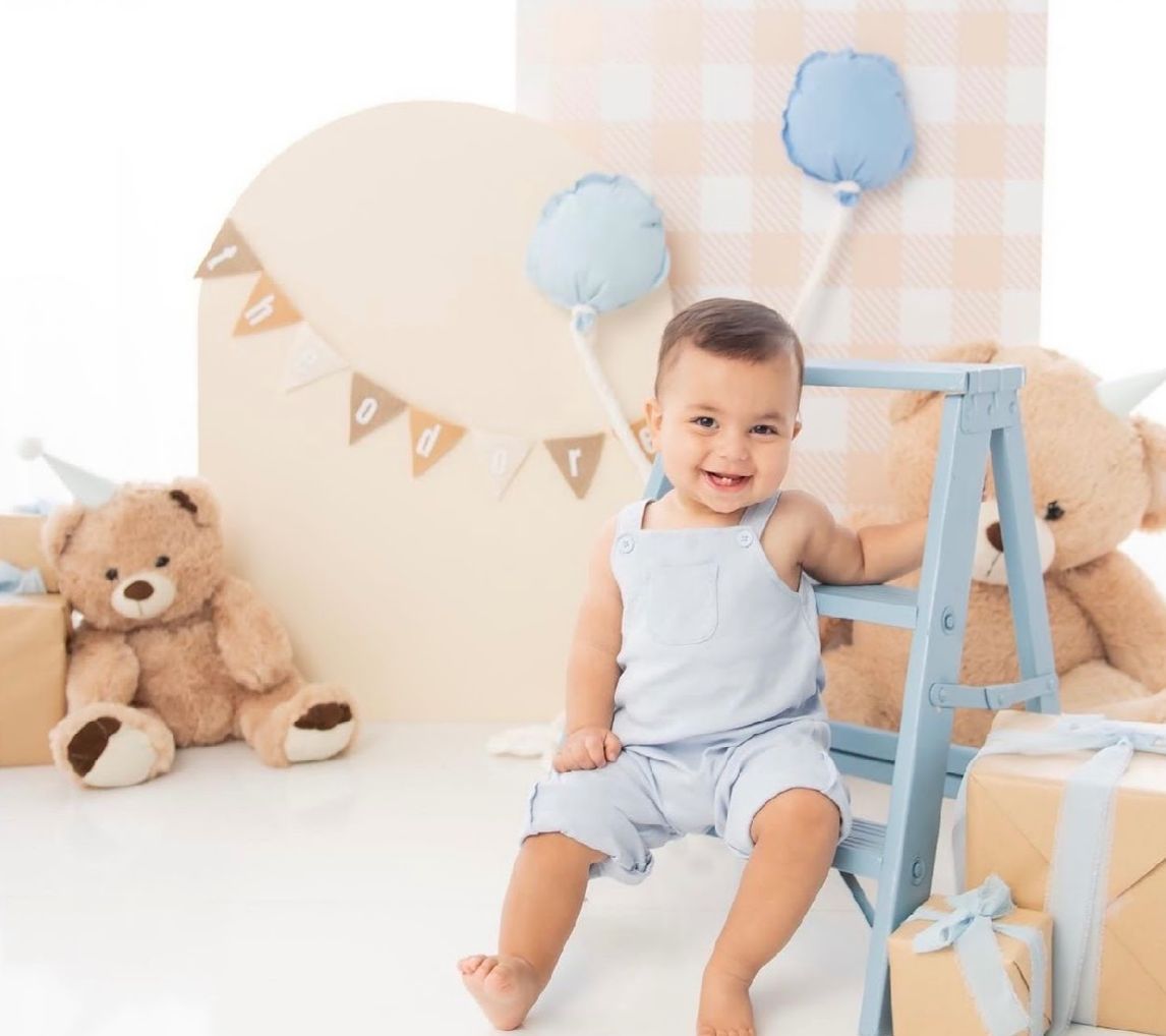 Baby in light blue outfit sits among teddy bears and pastel party decorations for a first birthday celebration.