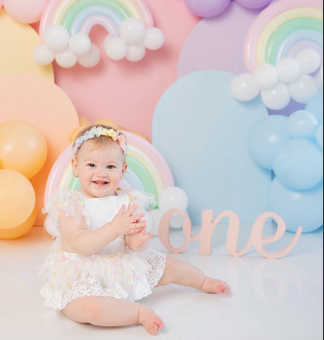 Baby in white lace dress sitting on pink backdrop with pastel rainbow and cloud decorations for cake smash photoshoot.