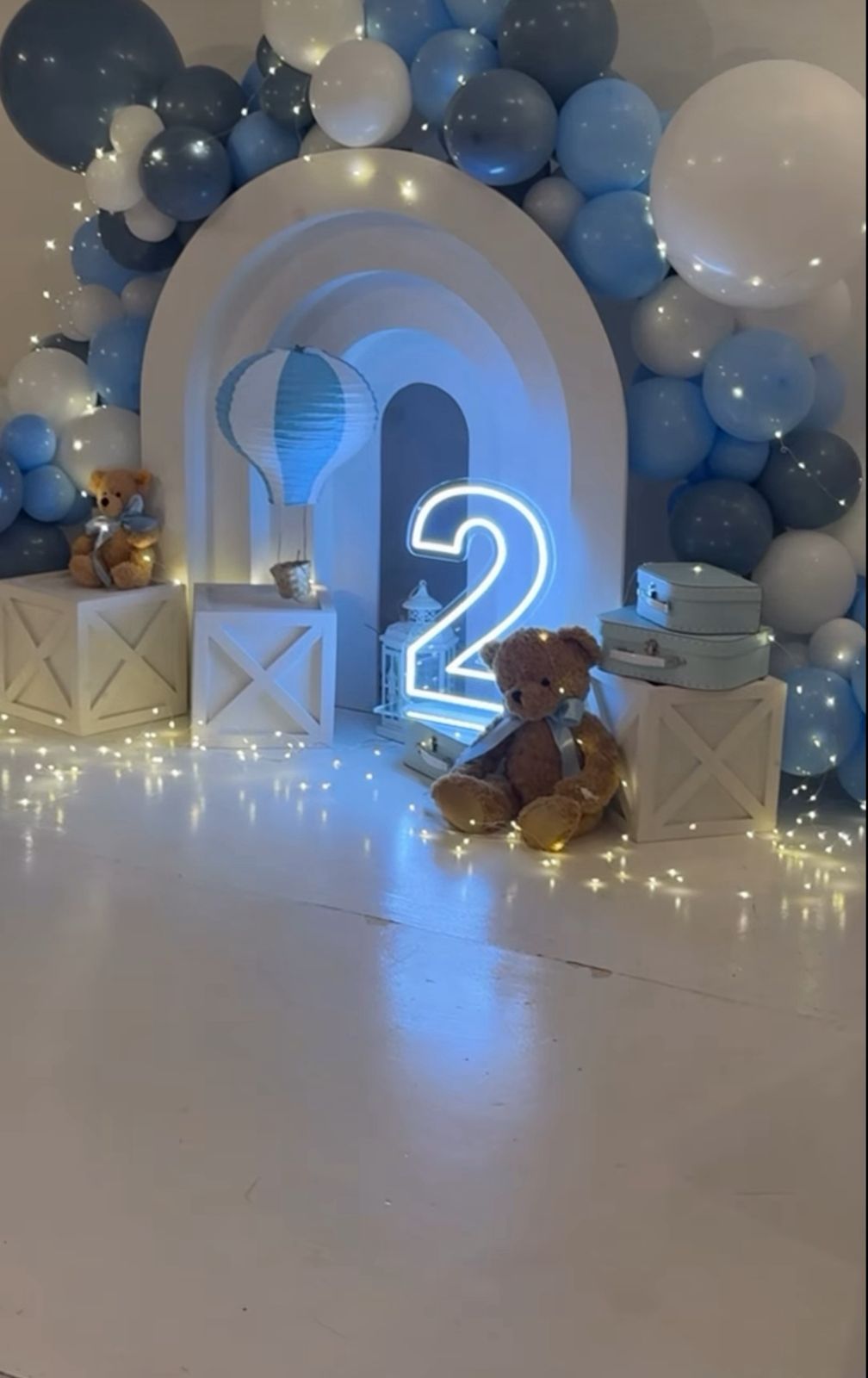 Blue and white balloon arch with neon number 2 sign and string lights creates a dreamy party atmosphere.