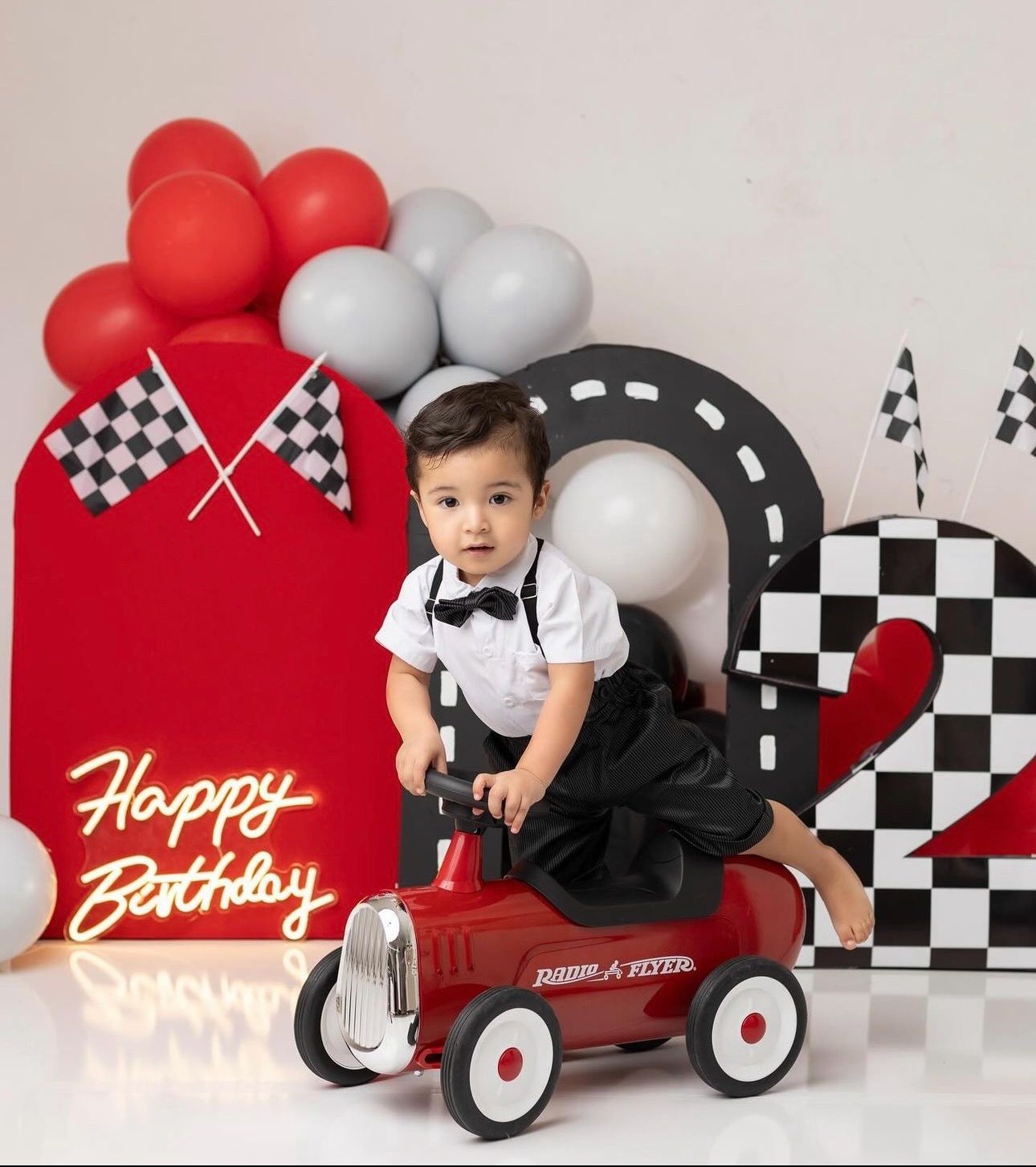Racing car themed birthday photo setup with red and white balloons, checkered pattern backdrop and vintage pedal car.