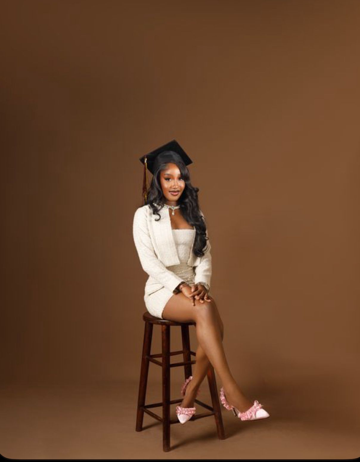 A figure in a white blazer and shorts sits elegantly on a wooden stool against a brown studio backdrop.