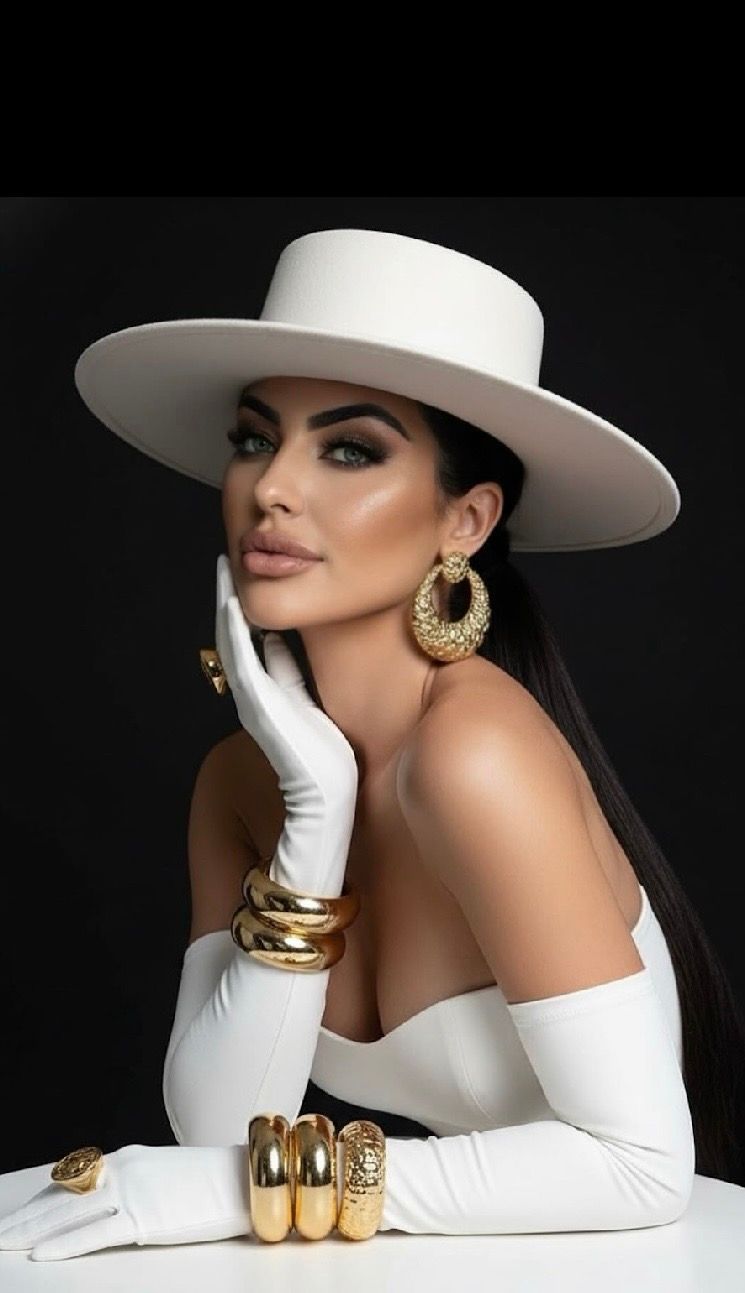 Woman in white wide-brim hat and gloves poses with gold jewelry including bangles and hoop earrings on dark background.