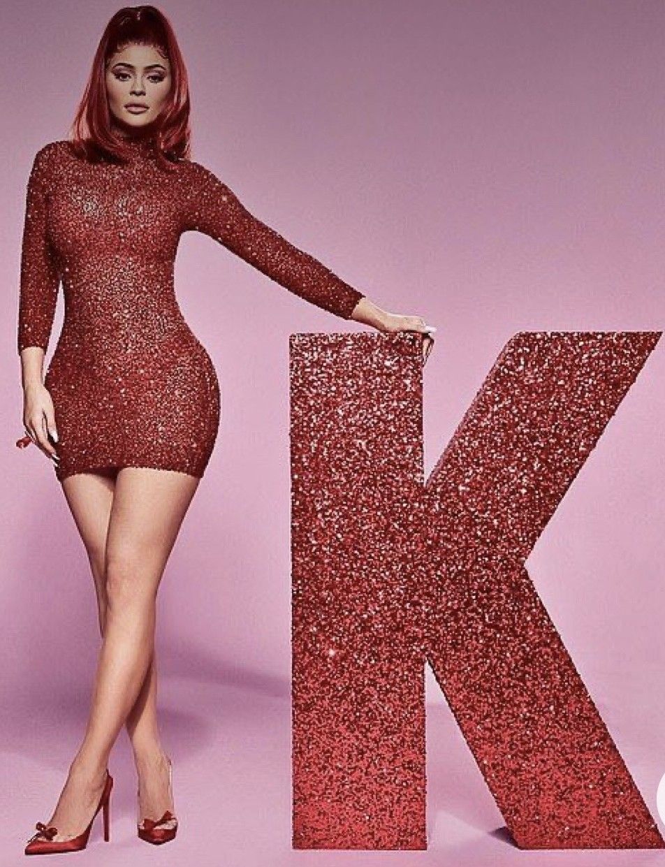 Woman in red sequin mini dress posing next to a glittery letter K on a pink background.