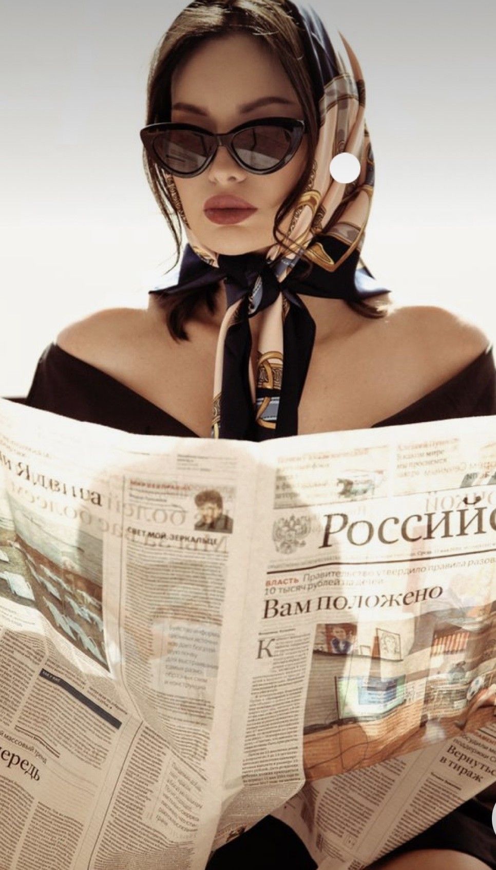 Stylish woman in cat-eye sunglasses and silk headscarf reading a Russian newspaper in a chic editorial pose.