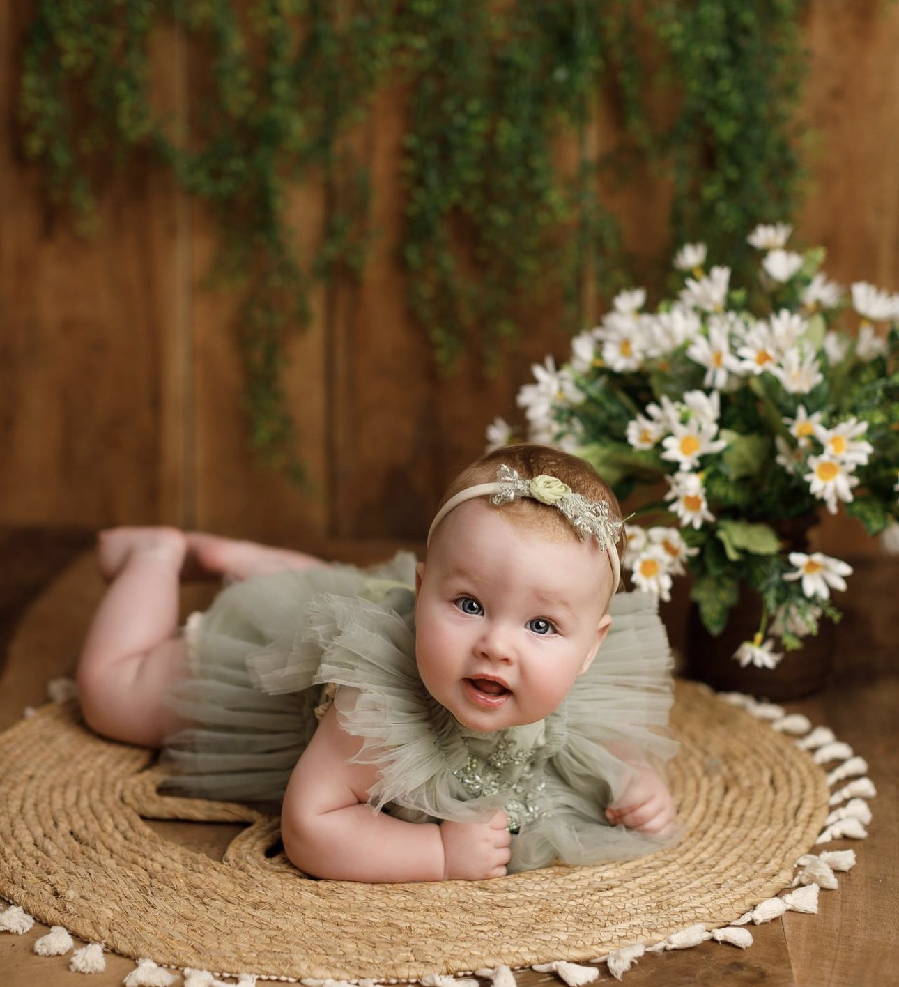 A baby girl in a sage green tulle romper and floral headband poses on a woven mat beside white daisy flowers.