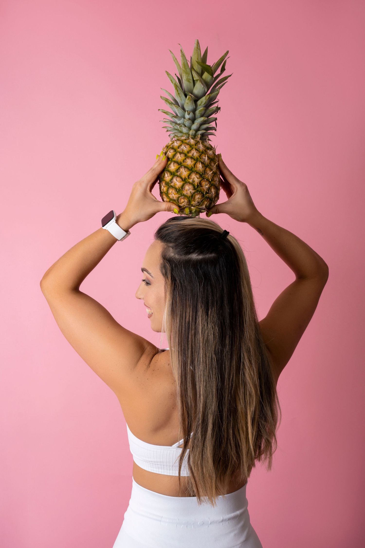 Athletic woman in white outfit holding pineapple overhead against pink background.