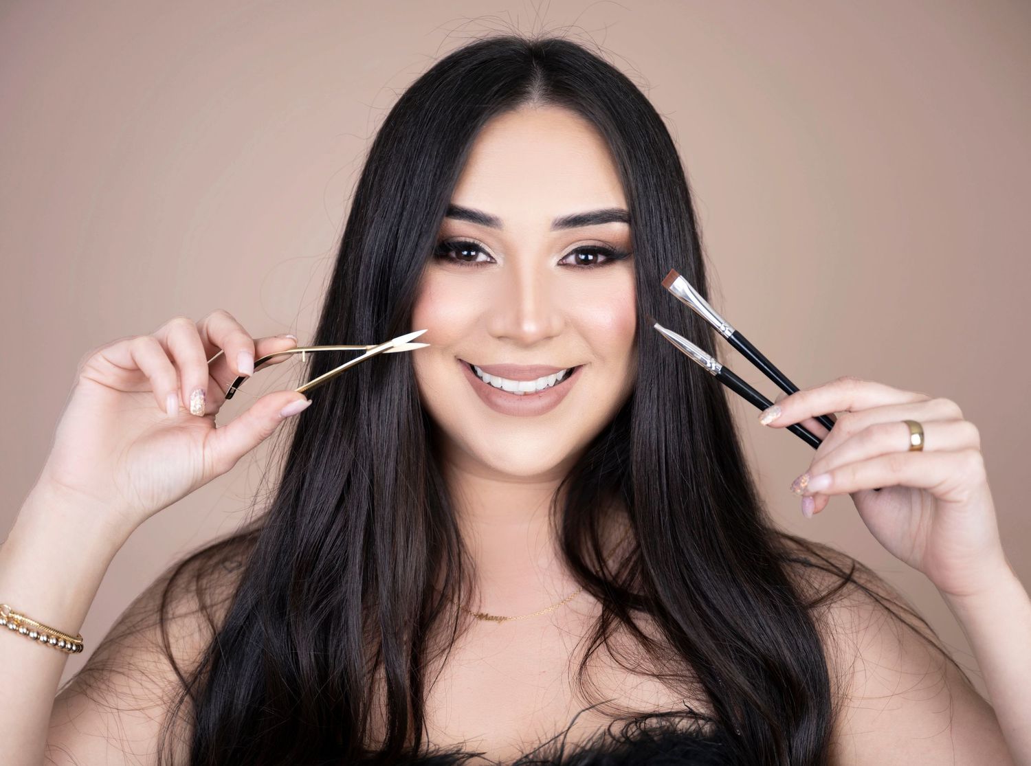 Woman with long dark hair holding makeup brushes near her face while smiling.