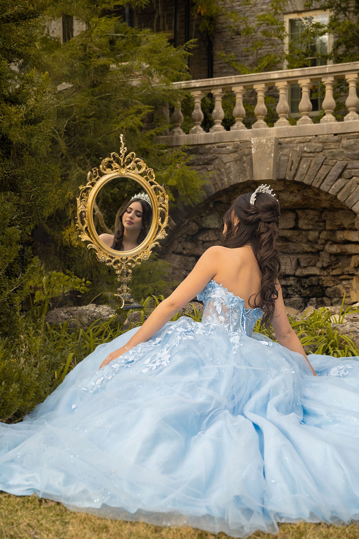 A young woman in a light blue ball gown and tiara sits before an ornate gold mirror in a castle garden.