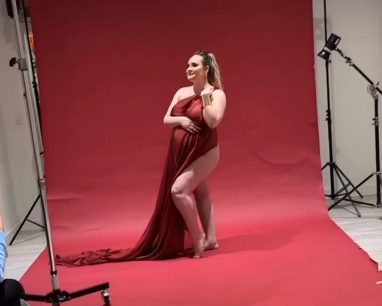 Pregnant woman posing in a flowing red maternity gown during a professional photo shoot against a red backdrop.