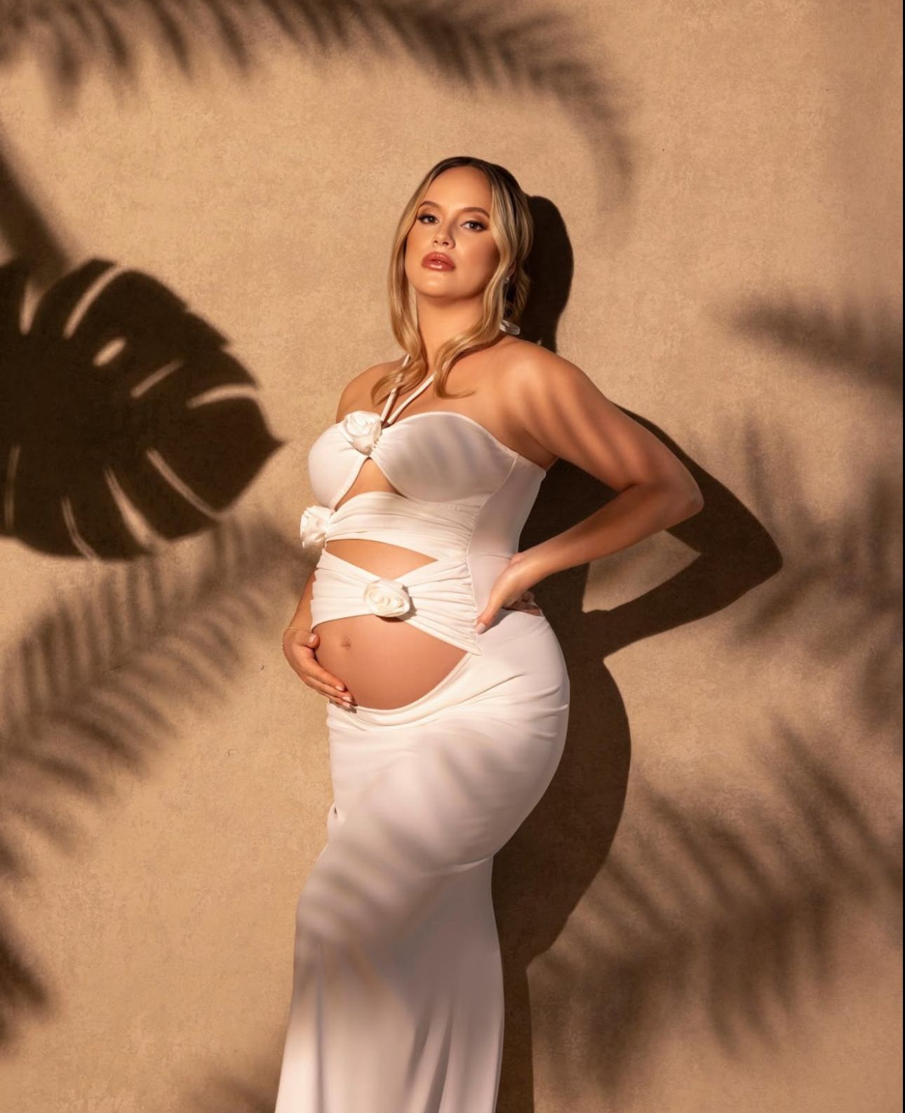 Pregnant woman in white cutout maternity gown posing with tropical leaf shadows on beige wall.