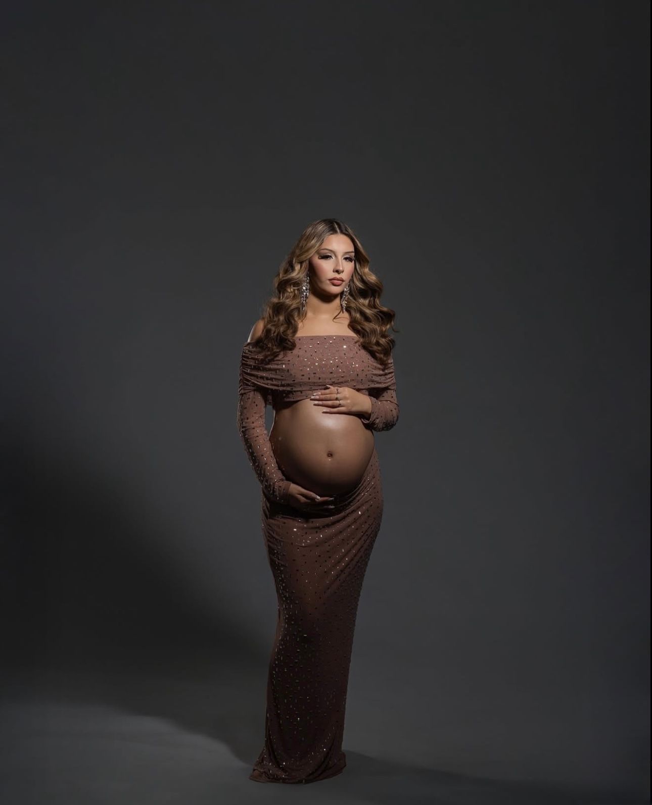 Pregnant woman in a sparkly mauve off-shoulder maternity gown posing elegantly against a gray studio background.