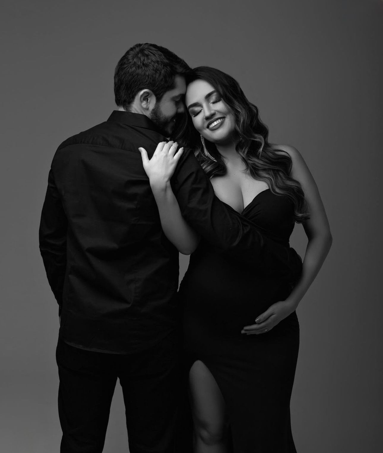 Black and white maternity photo of a couple in elegant black outfits, woman smiling as man whispers to her.