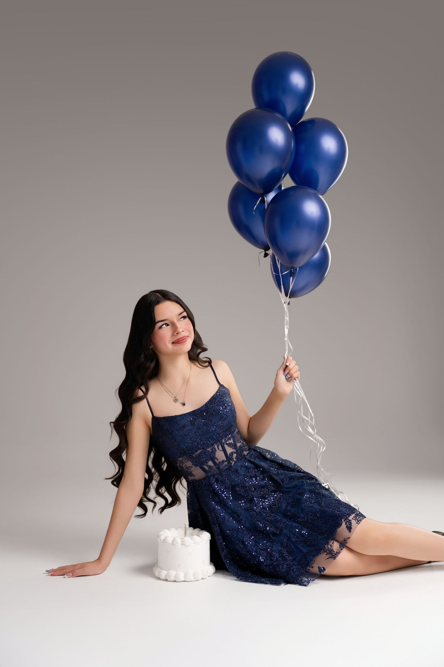 Young woman in blue lace dress sitting on floor holding blue balloons beside white cake on gray background.