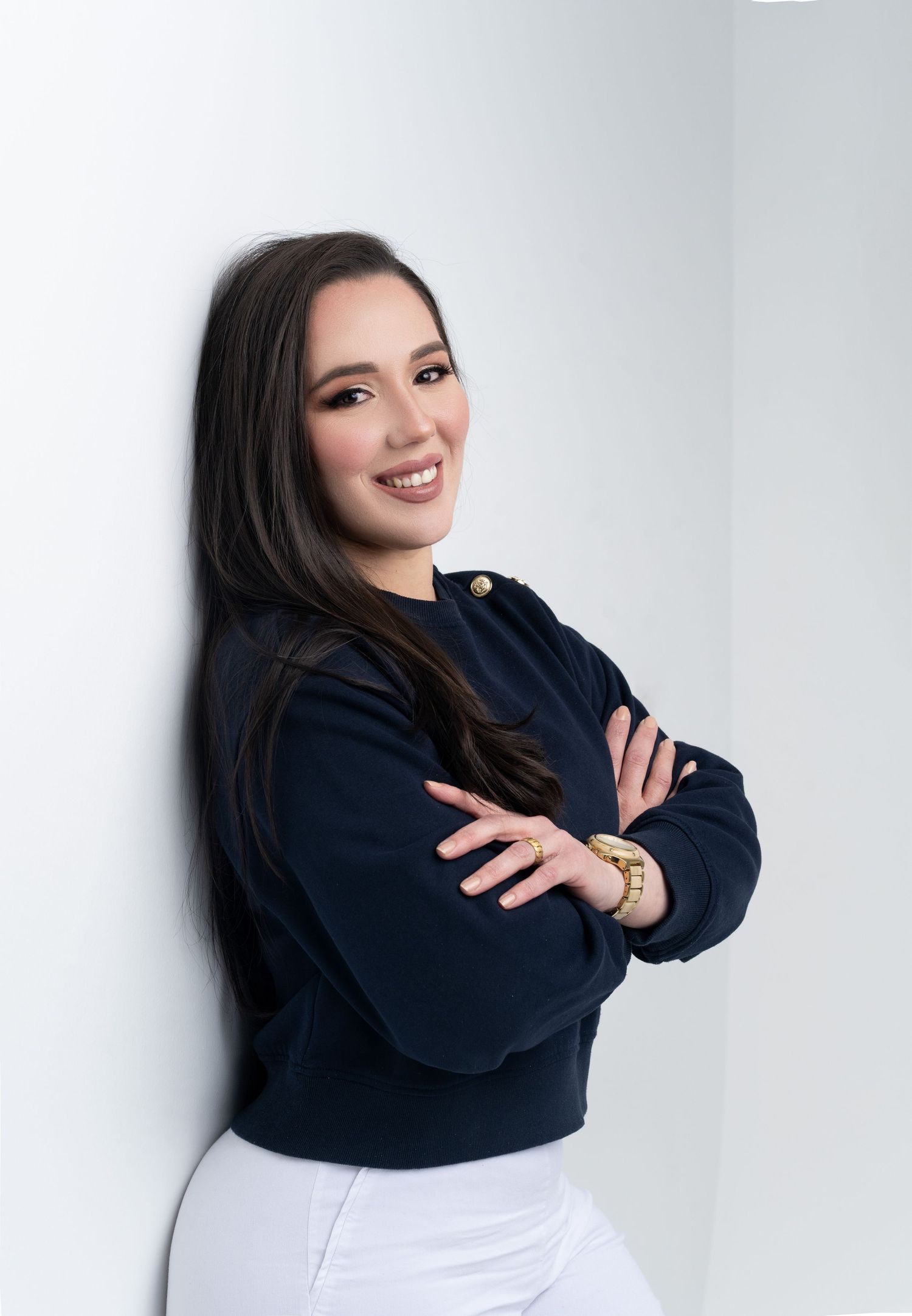 Smiling young woman with dark hair in navy sweater and white pants, arms crossed, leaning against white wall.