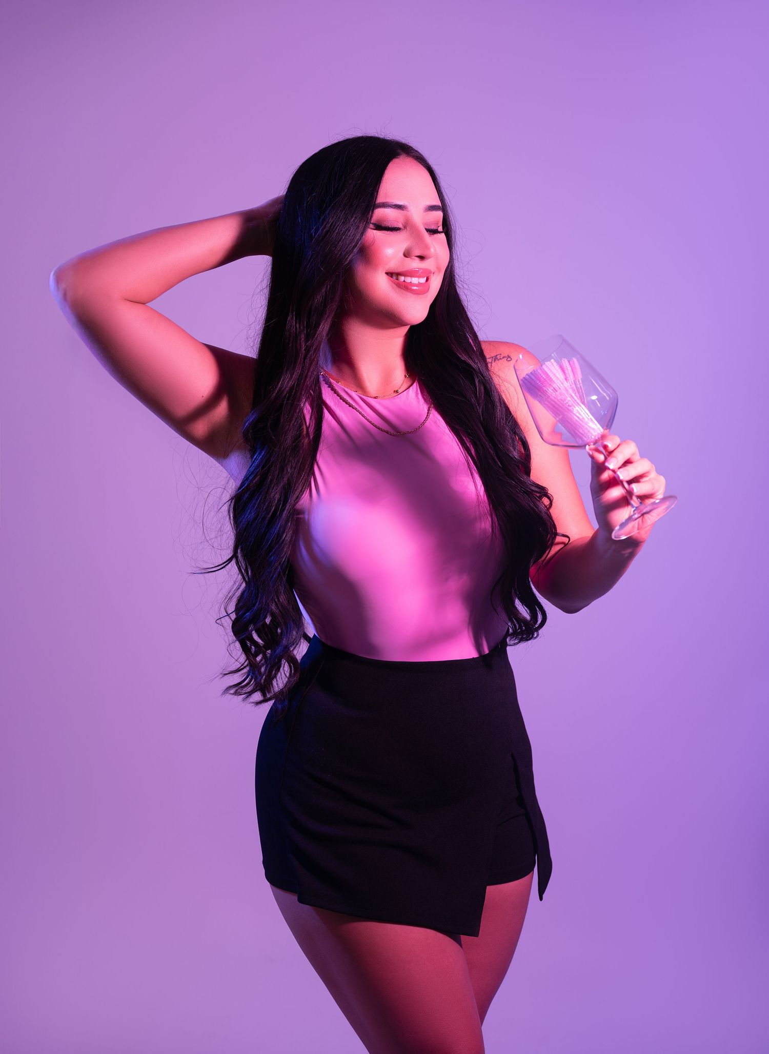 Smiling woman with long wavy black hair poses in a pink top and black shorts, holding a wine glass against a purple studio backdrop.