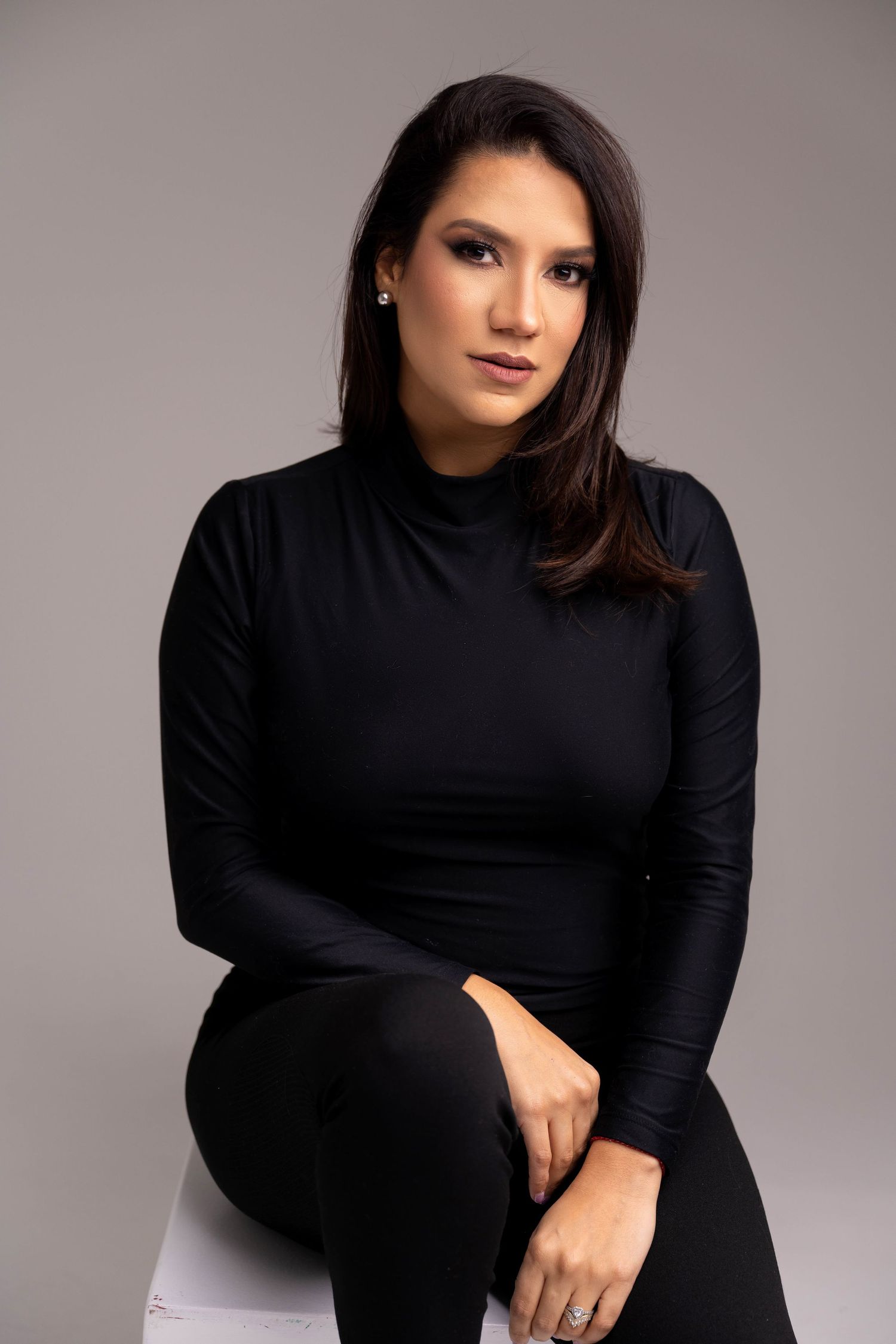 Woman with long dark hair in a black turtleneck and pants sits on a white stool, posing against a gray studio backdrop.