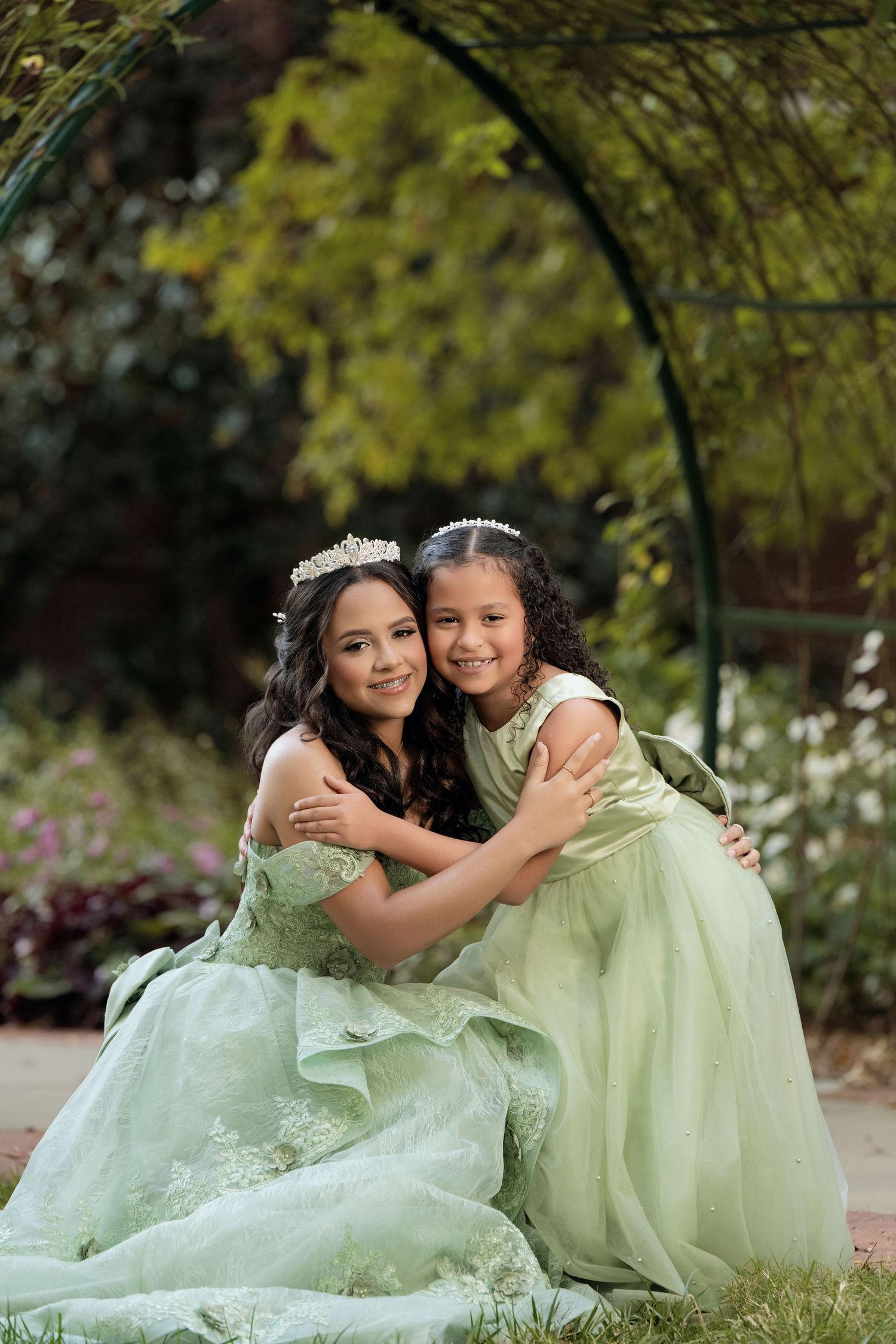 Two girls in matching sage green gowns and tiaras embrace in a garden setting.