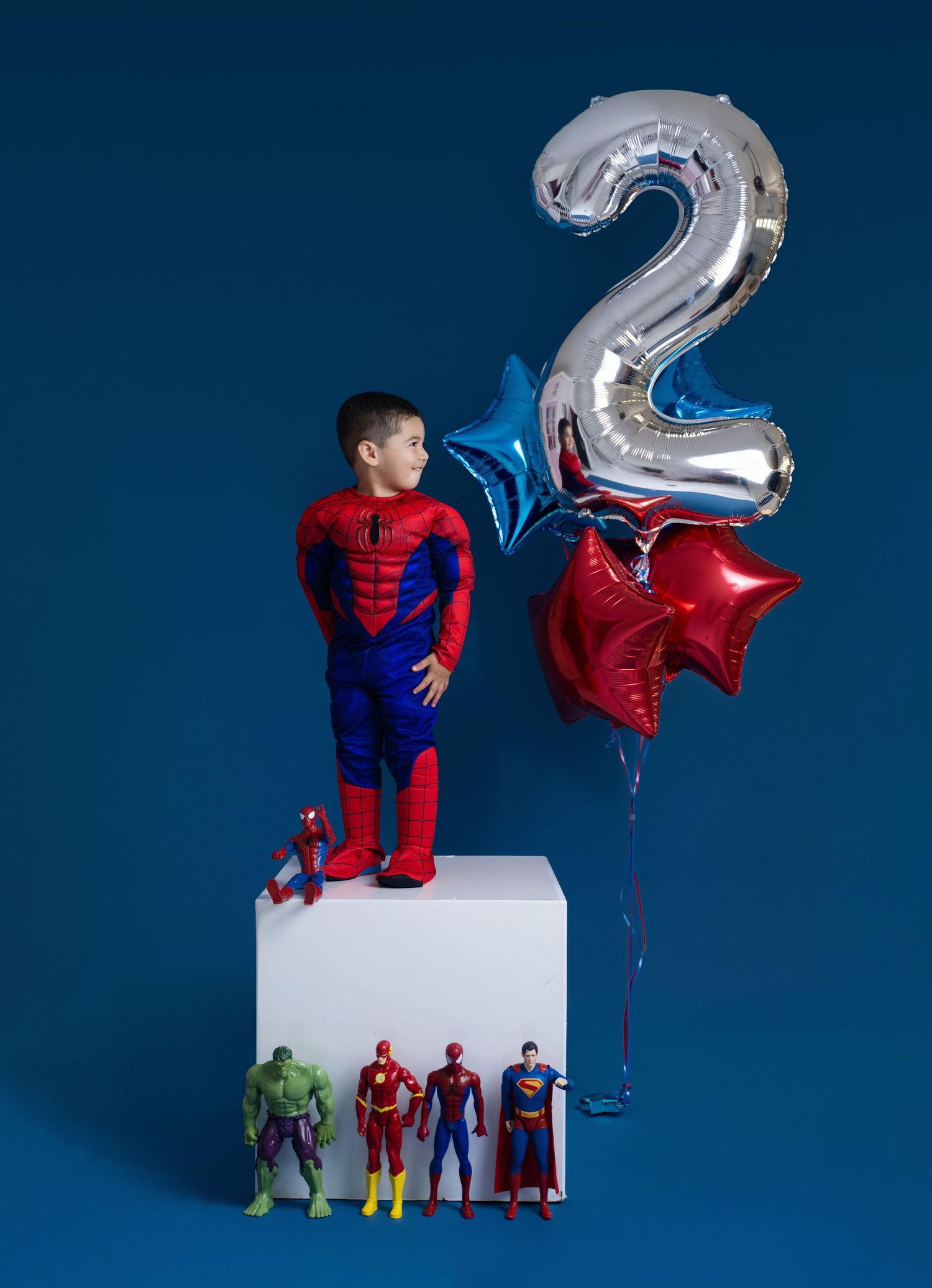 Boy in Spider-Man costume stands on white cube with superhero figures and a silver number 2 balloon for 2nd birthday.