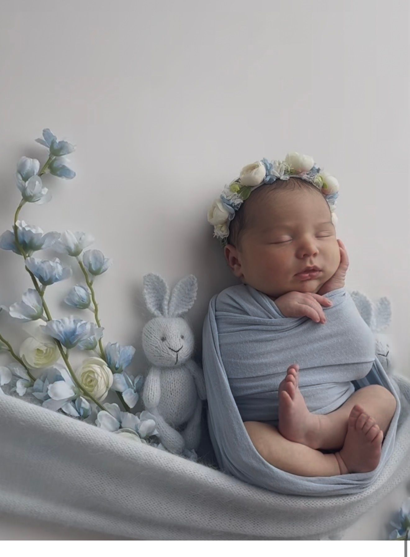 Sleeping newborn baby wrapped in blue cloth with floral crown, bunny toy, and blue flowers.