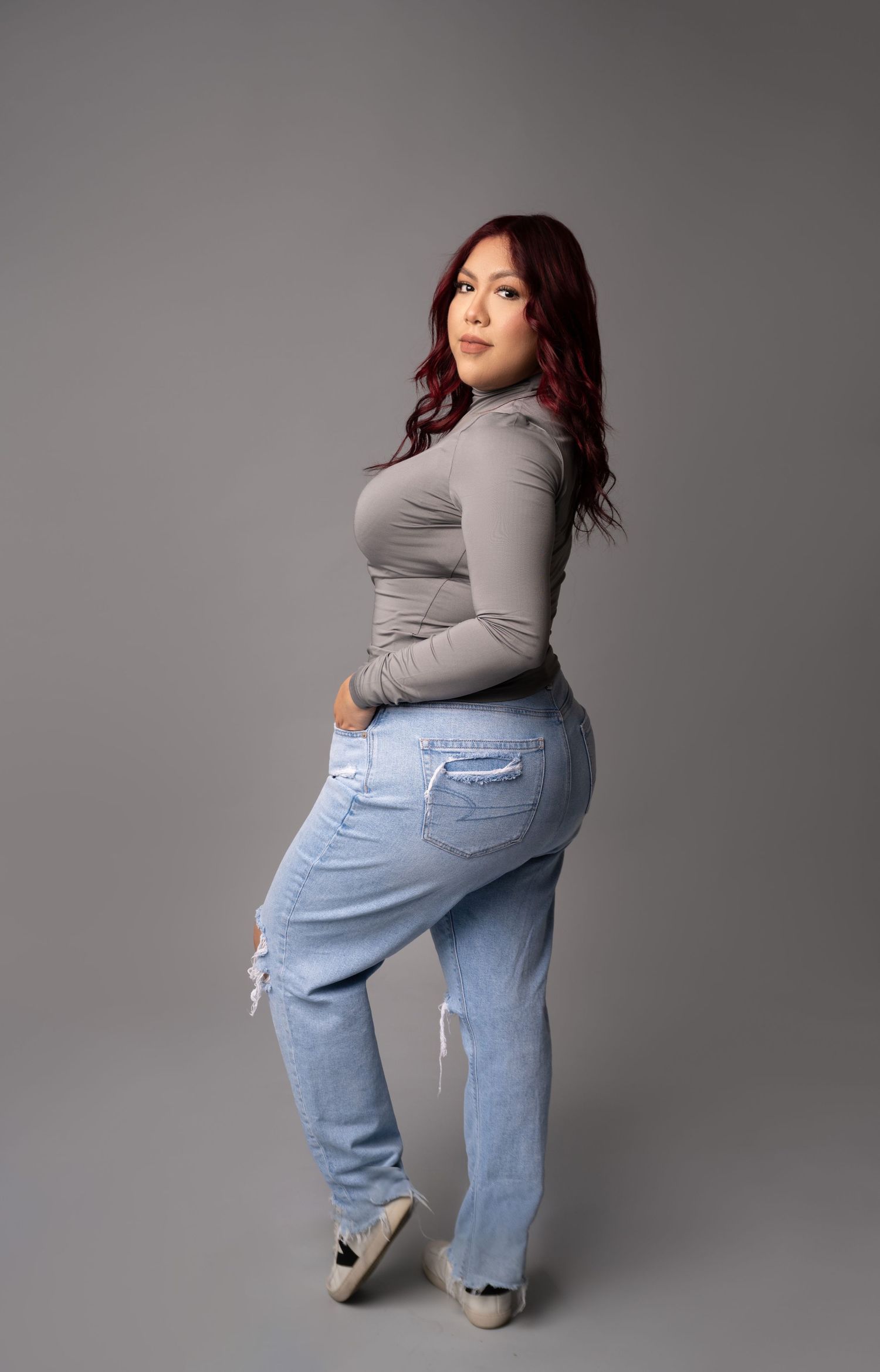 Woman with long burgundy hair poses in a gray long-sleeve top, light-wash ripped jeans and white sneakers against a gray studio backdrop.