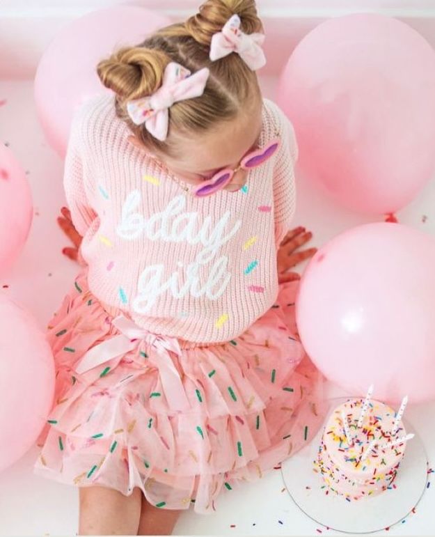 Young birthday girl wearing pink sprinkle tutu skirt and bday girl sweater, surrounded by pink balloons with cake.