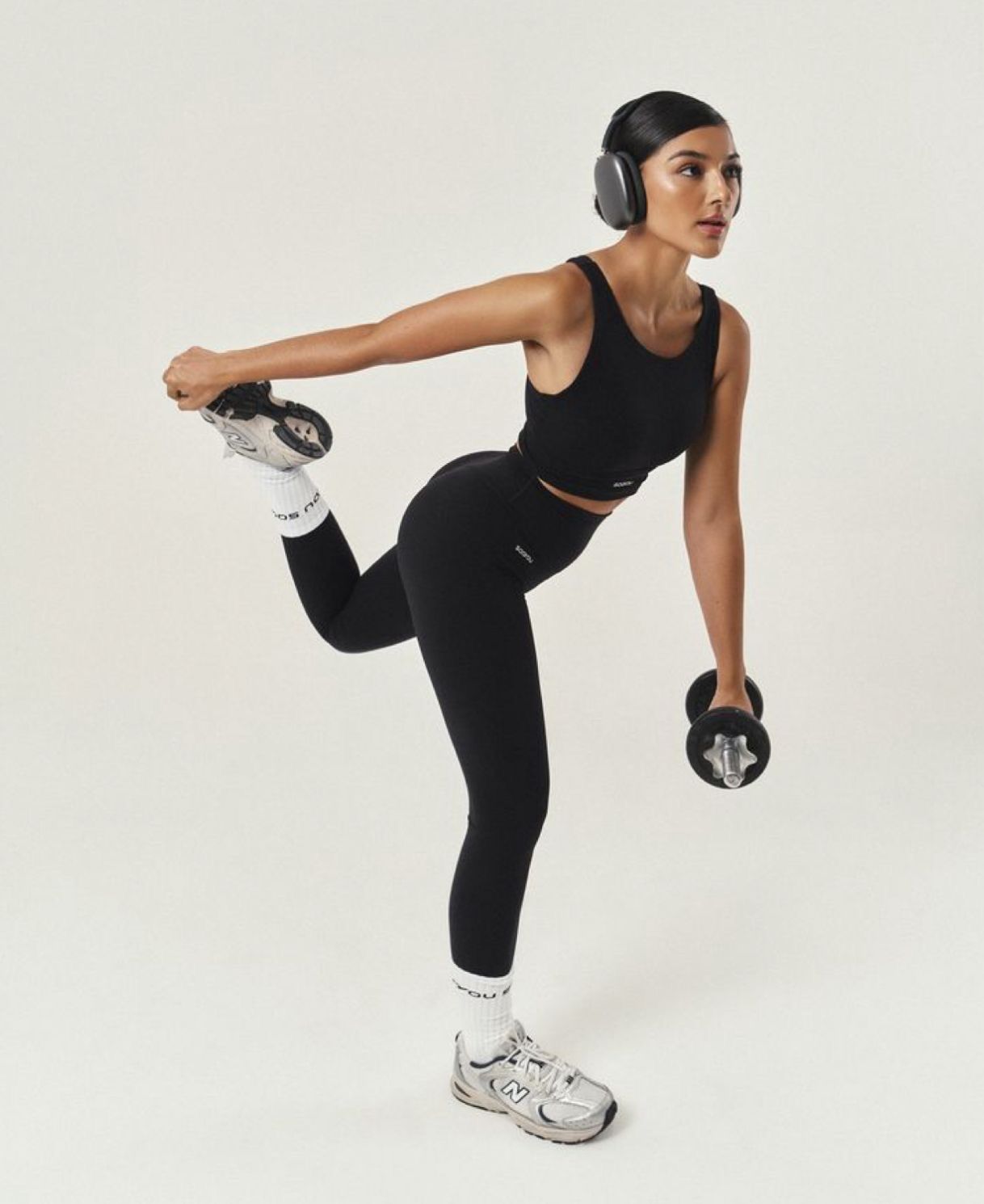 Woman in black athletic wear doing a single-leg balance exercise with dumbbells while wearing wireless headphones.