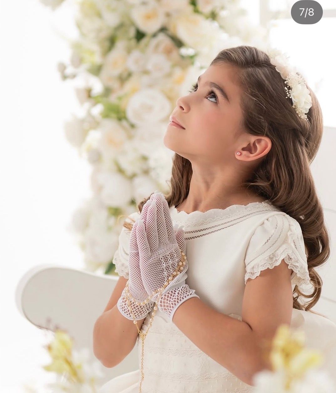 Young girl in white First Communion dress and lace gloves, hands clasped in prayer, looking upward among white flowers.