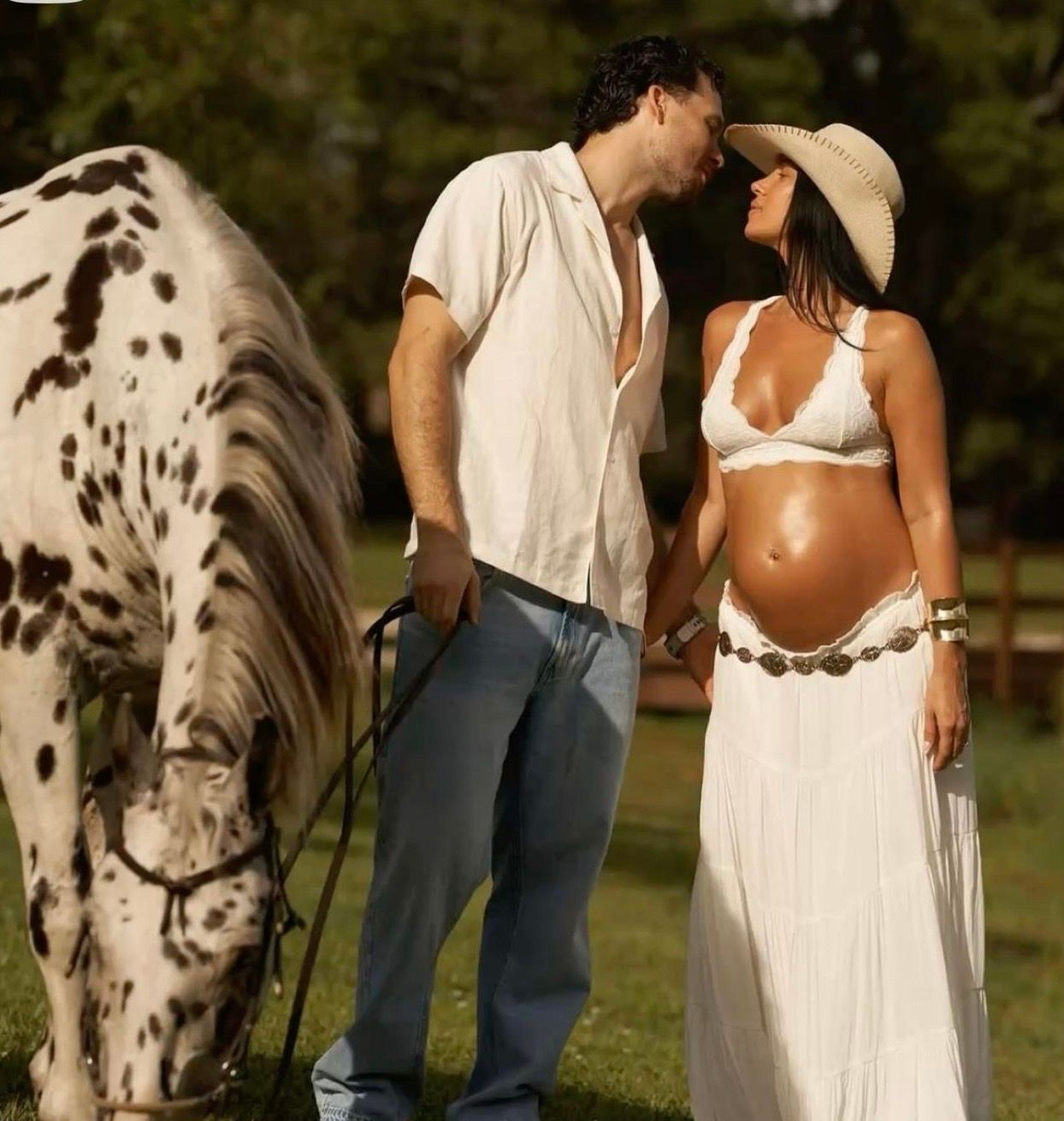 Pregnant woman in white outfit with man kissing near spotted horse on green field.
