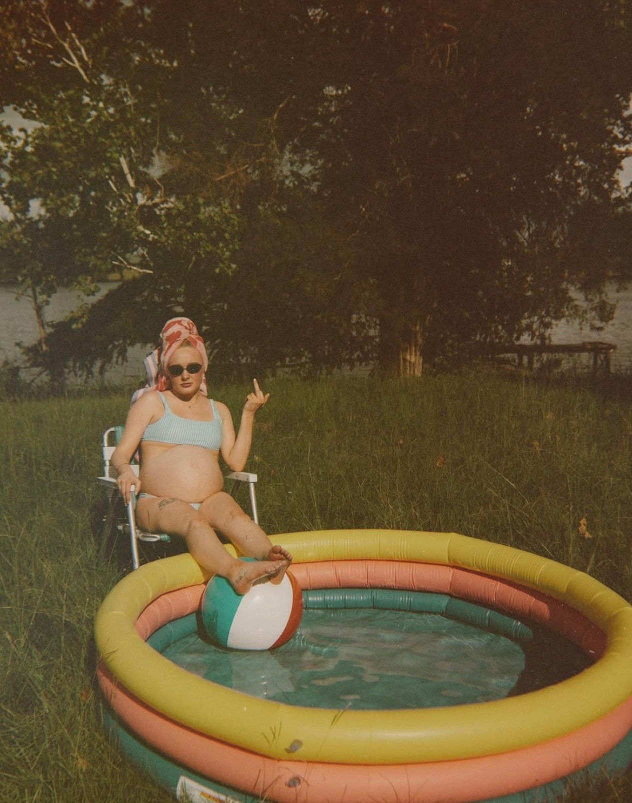 Pregnant woman in bikini sitting by a small inflatable kiddie pool in a backyard, holding a beach ball.