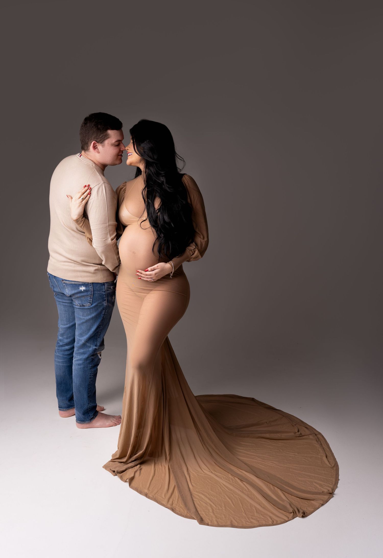 A couple in casual attire dances gracefully together against a gray studio backdrop, her tan dress flowing elegantly.