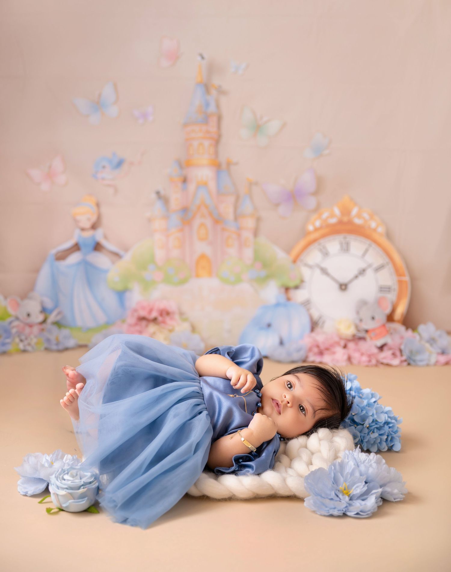 Baby girl in blue dress poses on white cloud prop with Cinderella castle backdrop and floral decorations.