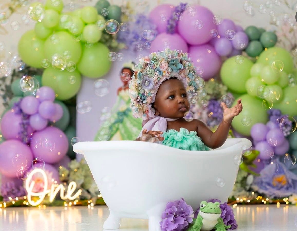 Baby girl in floral bonnet sits in white tub at Princess Tiana themed first birthday party with balloon arch.