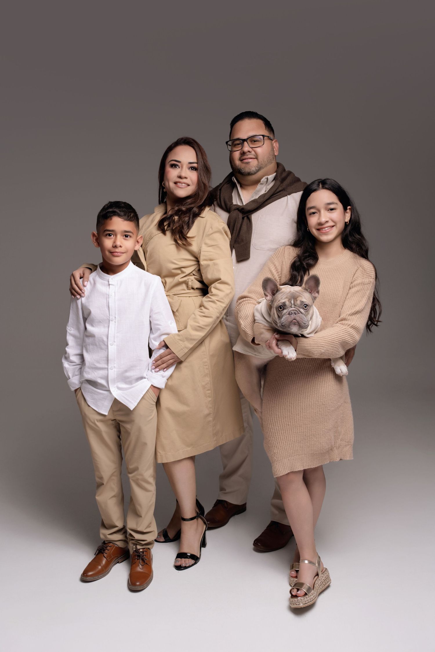 Family of four posing with a French Bulldog in matching beige outfits against a gray studio background.