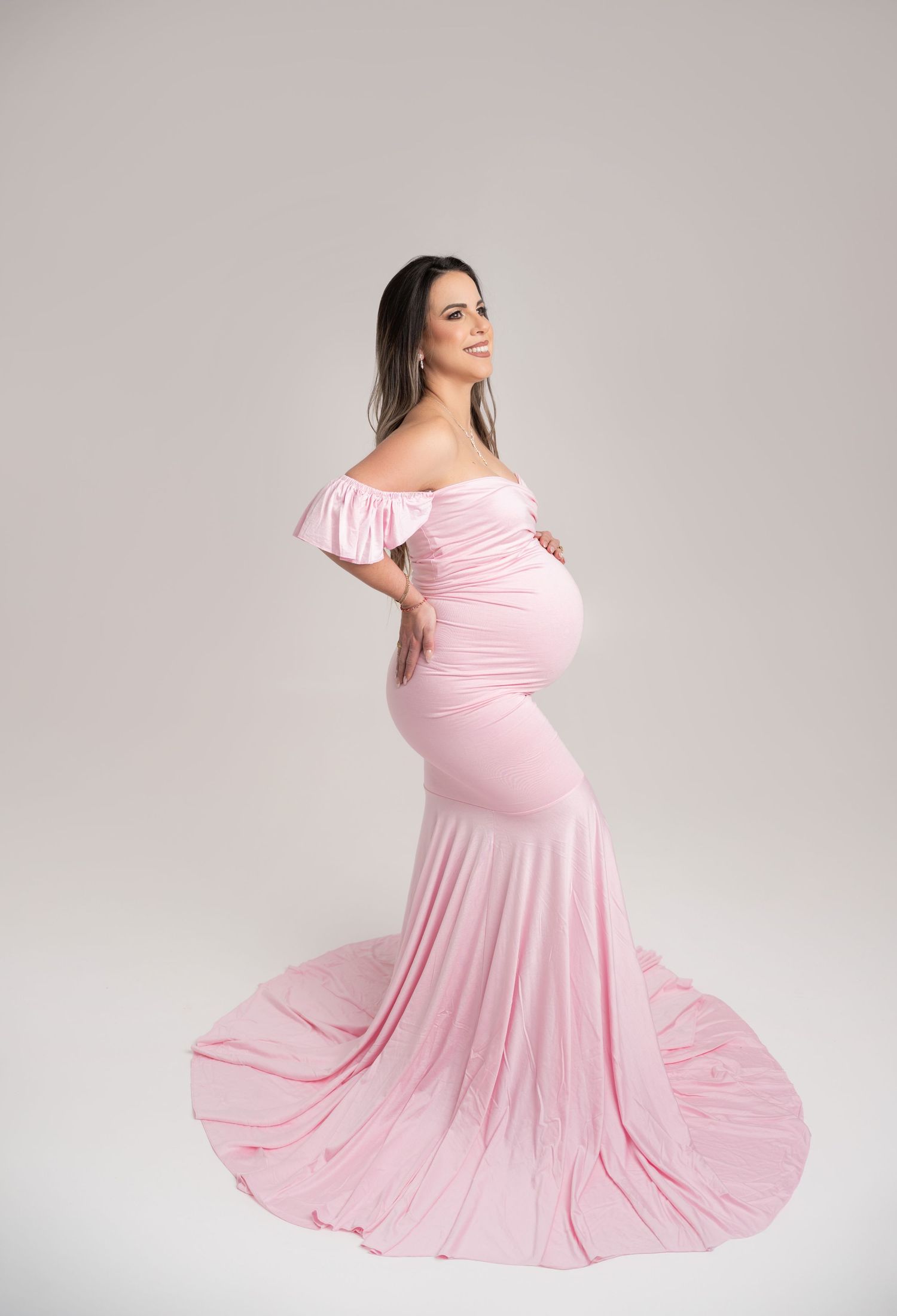 Pregnant woman in elegant off-shoulder pink mermaid maternity gown posing gracefully in a studio setting.