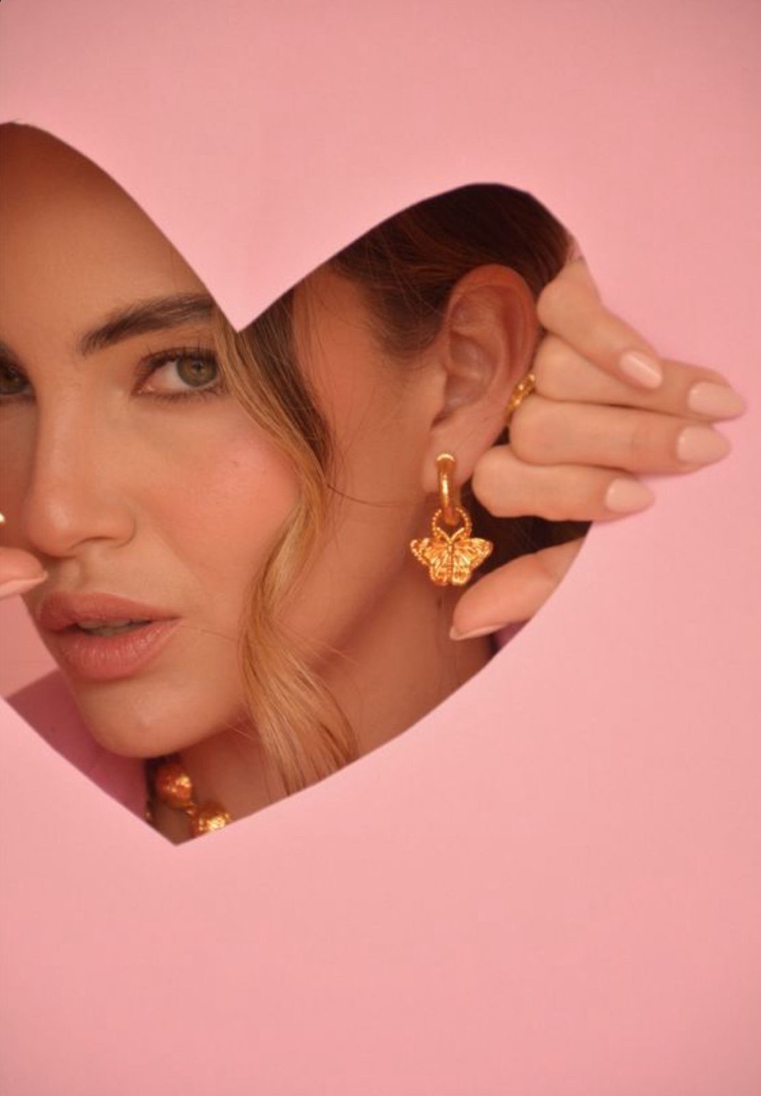 Woman wearing gold floral charm hoop earrings, viewed through a heart-shaped cutout in pink paper.