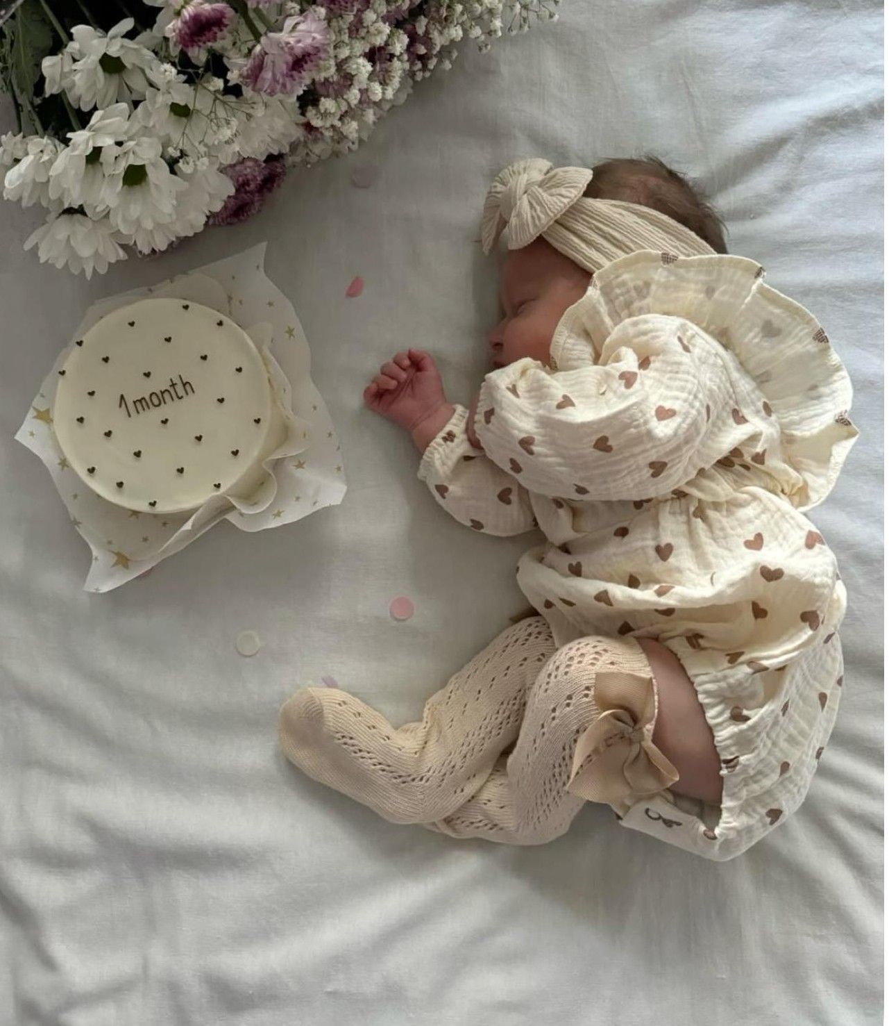 A newborn baby in a cream heart-print outfit lies next to a 1-month milestone card and fresh flowers on white bedding.