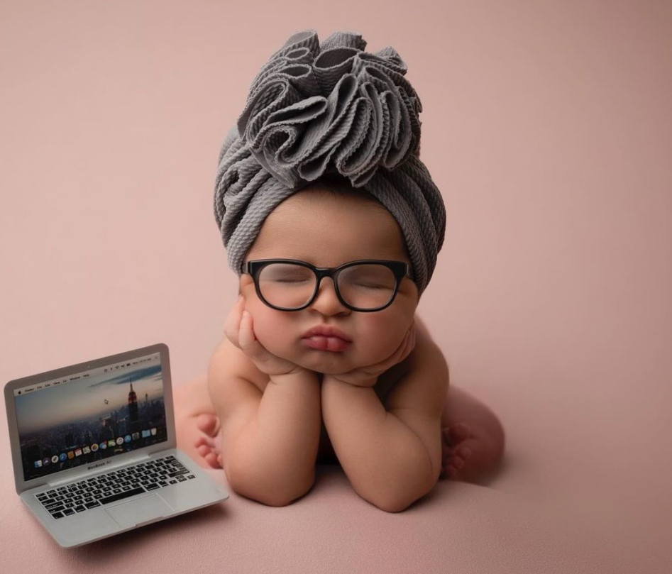 A baby wearing glasses and a gray turban poses next to a laptop computer on a pink background.