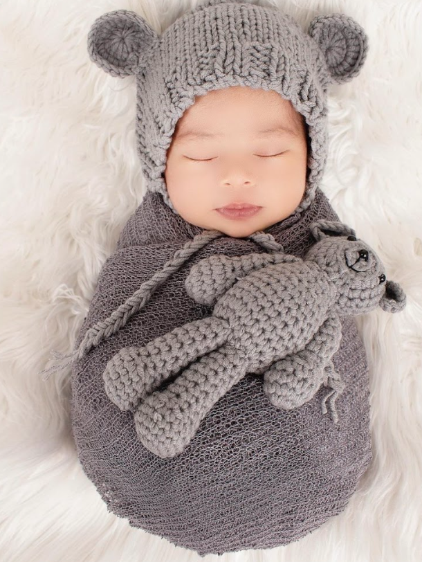 Sleeping baby wrapped in gray knit blanket with matching bear hat and stuffed animal on white fur background.