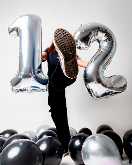 A person stands among black and silver balloons with large silver number balloons showing '12' against a white background.