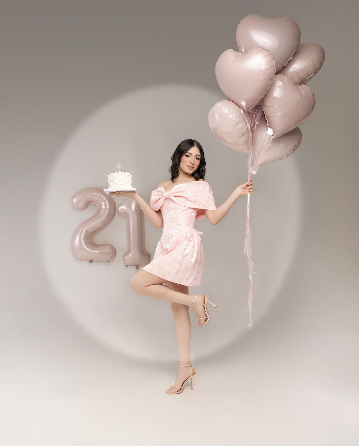 A person in a pink dress poses gracefully with rose gold heart balloons against a light gray studio backdrop.