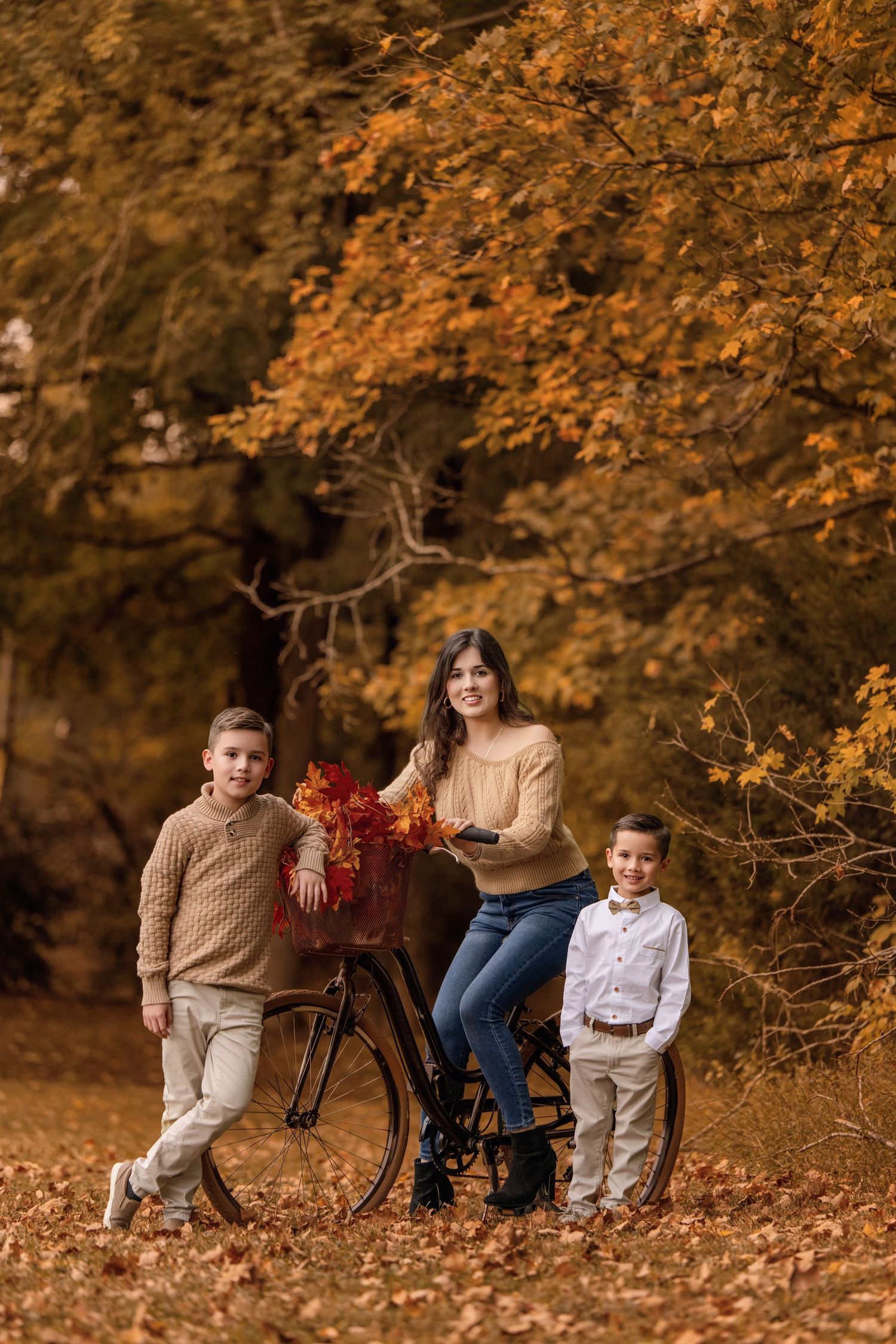A mother sits on a vintage bicycle with autumn leaves in the basket, flanked by her two young sons in a fall forest.