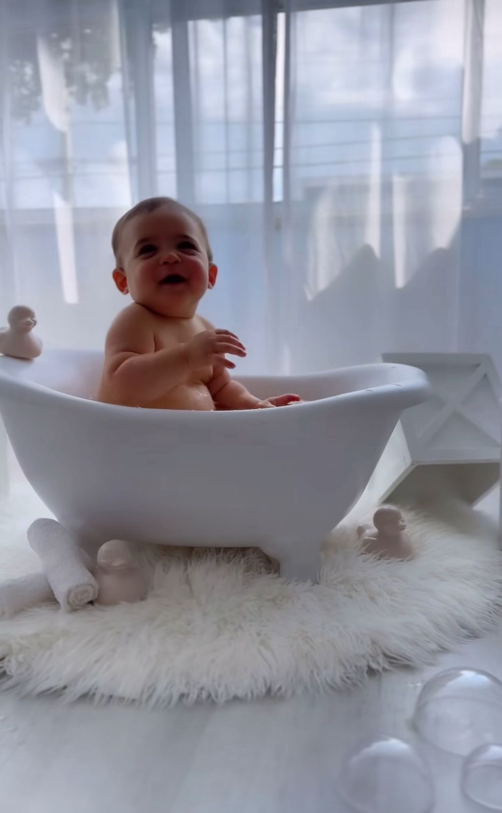 Happy baby enjoying bath time in white tub placed on fluffy white rug.