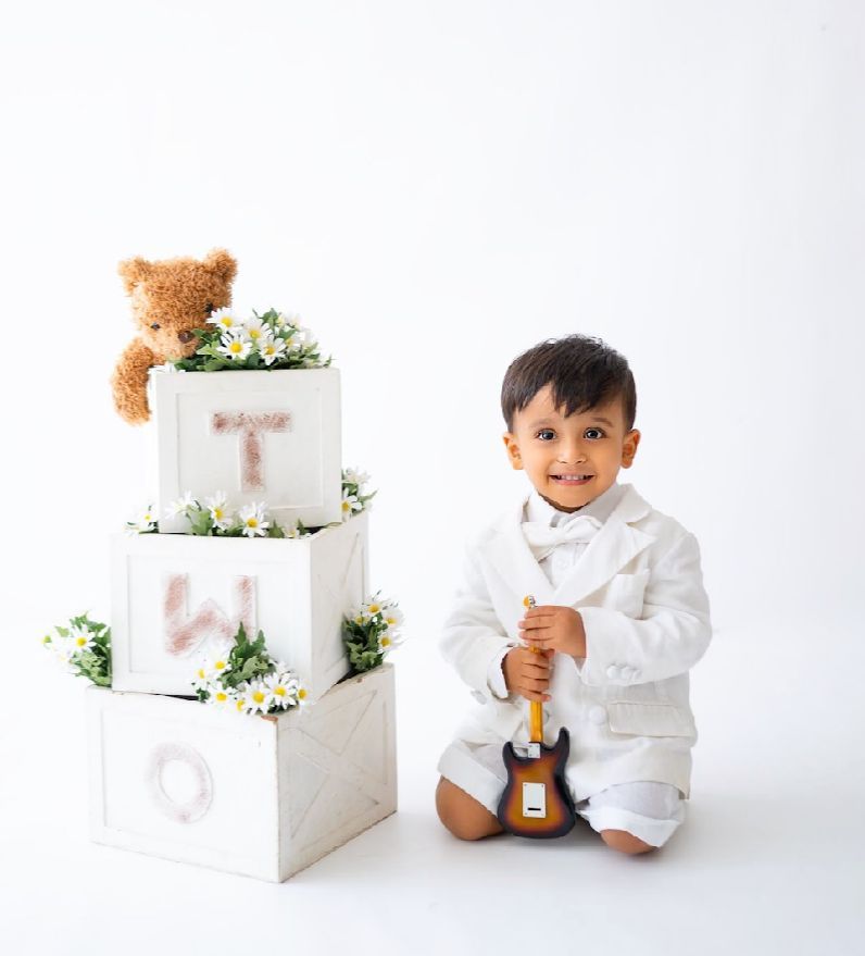 A baby in white clothing sits near elegant white tiered boxes decorated with delicate floral arrangements.